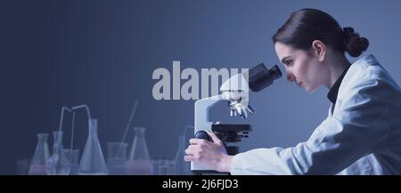 Young researcher working in the lab, she is looking through a microscope and checking samples, medical research concept Stock Photo