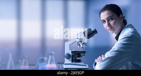 Young female researcher using a microscope and looking at camera, science and medical research concept Stock Photo