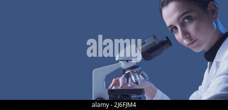 Young female researcher using a microscope and looking at camera, science and medical research concept Stock Photo