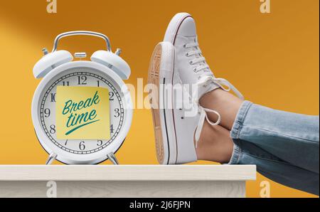 Woman with feet up on a desk and alarm clock with sticky note, it's break time Stock Photo