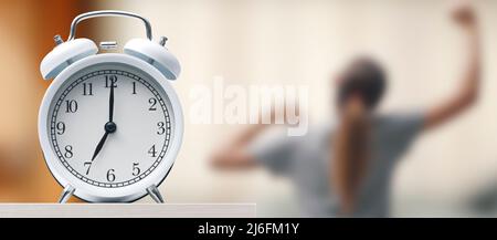 Alarm clock on the bedside table, a woman is getting up and stretching Stock Photo