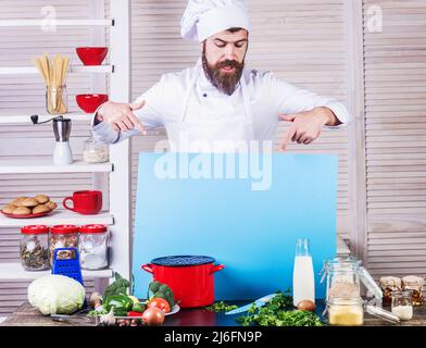 Male chef in uniform pointing with fingers at blank menu board. Cooking food. Professional kitchen. Stock Photo