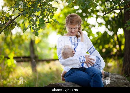 Mother breastfeeding baby son outdoors. Woman and her son wearing Ukrainian style shirts while sitting under tree Stock Photo