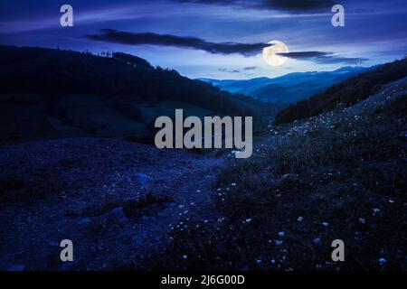 mountainous countryside landscape at night. rural fields on the hills. village in the distant valley. gorgeous sky with clouds glowing in full moon li Stock Photo