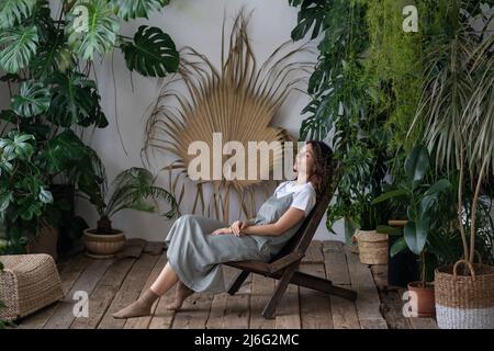 Young woman relaxed sit on comfortable armchair in stylish indoor garden with green exotic plants Stock Photo