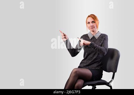 A girl, in business clothes, sits on a chair and indicates the direction with her hands, on a gray background. High quality photo Stock Photo