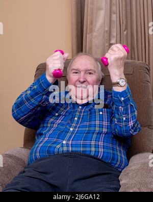 smiling man exercises with small barbells from the comfort of his living room chair Stock Photo