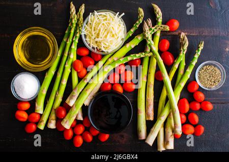 Caprese Roasted Asparagus Ingredients on a Dark Wood Table: Fresh asparagus, grape tomatoes, and other ingredients on a dark background Stock Photo