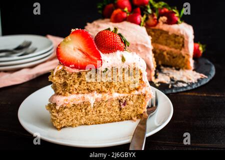 Slice of Fresh Strawberry Cake on a White Plate: A slice of cake on a small plate with the rest of the cake in the background Stock Photo