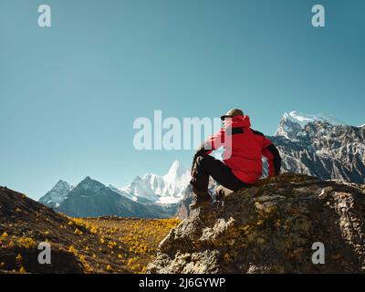 asian man sitting on top of rock with Yangmaiyong (or Jampayang in Tibetan) mountain peak in the distance in Yading, Daocheng County, Sichuan Province Stock Photo