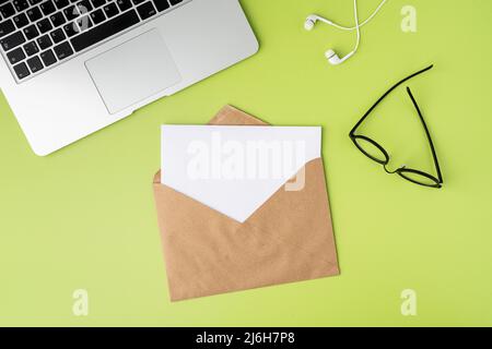 Concept of open craft envelope with blank paper sheet on green background. Laptop, earphones, glasses Stock Photo