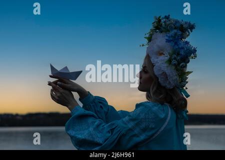 Young beautiful Ukrainian woman in a blue embroidered shirt, holding a paper boat made of white paper and looking forward with hope. Stock Photo