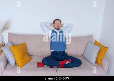 Successful young business man sits on sofa in lotus position in blue suit with notepad and thinks about business plans. Caucasian businessman meditates in the office on the couch Stock Photo