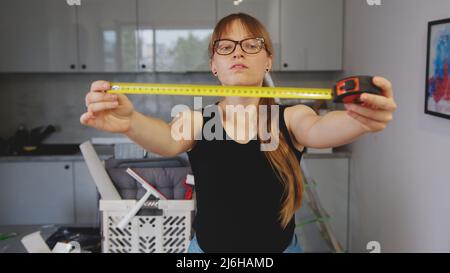 Indoor portrait of confident focused red-haired woman with tape measure in hands showing it to the camera while standing on background of workshop. High quality photo Stock Photo