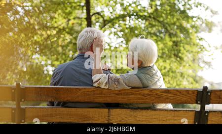 Outdoor photo of senior caucasian heterosexual couple sitting on a bench in the park enjoying beautiful weather. Gray-haired lady in warm vest touches her husband's hair. High quality photo Stock Photo