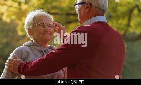 Beautiful relationship. Elderly caucasian heterosexual couple being affectionate. Gray-haired man in red sweater touches glasses of his partner. High quality photo Stock Photo