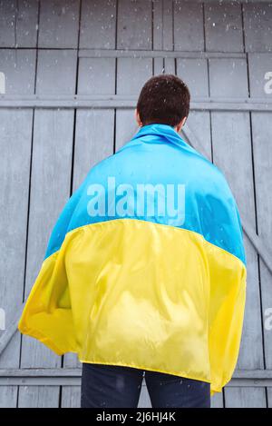 The man is wearing the state flag of Ukraine on his shoulder and is standing with his back to the camera outdoors in snowy weather. Ukrainian flag, patriotism. Independence Flag Day of Ukraine. Stock Photo