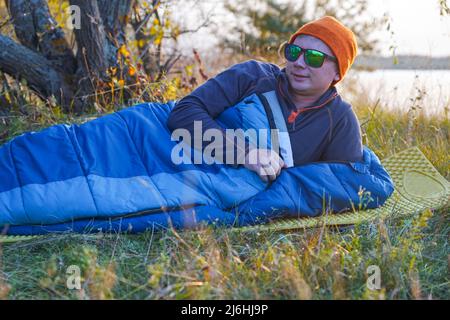 Guy tourist lies in a sleeping bag on the bank of the river. Nearby are his things: a backpack, boots, a thermos, a rug. Stock Photo