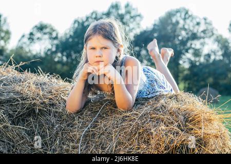 Portrait of little blond girl in dress alone lying and relaxing on haystack in field. Light sunny day. Rest and calm concept. Fresh air in countryside Stock Photo