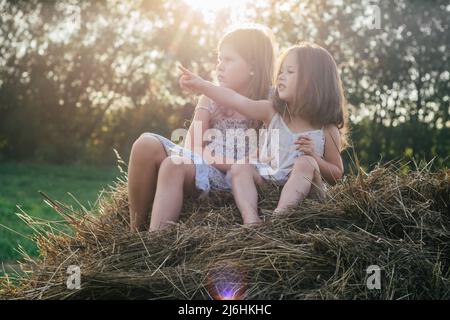 Portrait of two children girls sitting on straw bale in hay field, point and look in same direction. Light sunny day. Cheerful and enjoy friendship Stock Photo