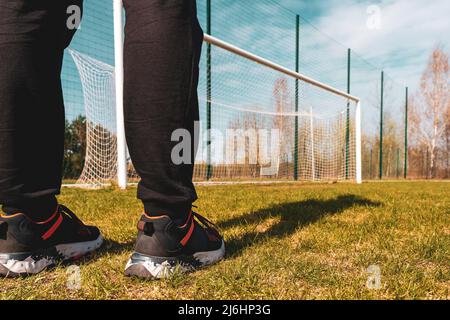 Low angle view of the feet of a sportsman who stands in a soccer field against a blue sky and the gate at the stadium. The game at the stadium. Stock Photo
