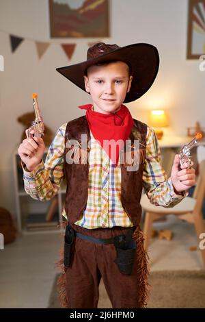 Happy cute little boy in costume of cowboy posing for commercial with two revolvers while standing in his room and looking at camera Stock Photo