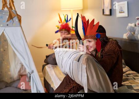 Happy cute girl in headdress of native Americans shooting from bow while sitting on bed during play with her friend or brother Stock Photo
