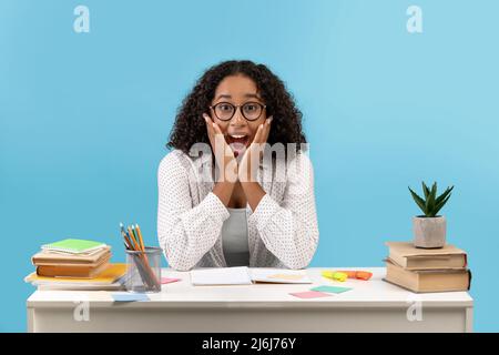 Excited young black woman in glasses sitting at desk with study materials, achieving success in exams on blue background Stock Photo