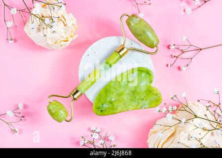 Green quartz stone gua sha scraper and jade roller for facial beauty lifting anti age massage with white flowers on pastel pink background top view. Stock Photo