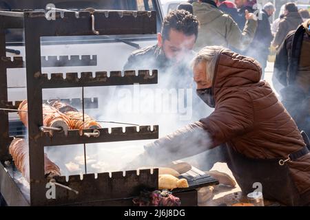 A street vendor preparing grilled lamb Kokorec on skewers over charcoal fire at a street festival for a customer. Covid-19 measure use face mask Stock Photo