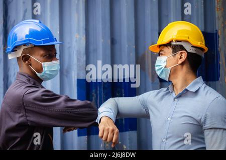african engineer and caucasian businessman wearing face mask to protect from covid or coronavirus in cargo warehouse at the dock have greeting and tou Stock Photo