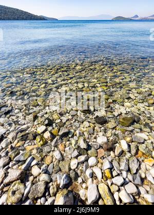Close-up of wet stones and sea. Pebble beach. Tiny rocks in crystal clear water. Horizon over water on sunny day. Sky reflecting on waves. Vertical Stock Photo