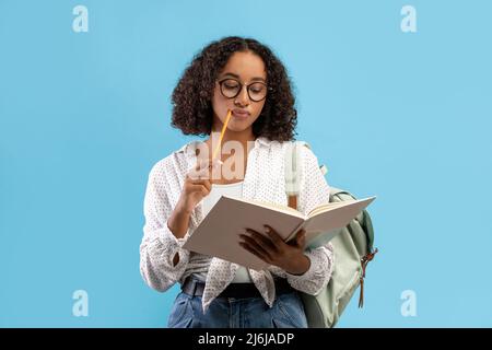 Thoughtful African American female student reading book, thinking over home assignment on blue studio background Stock Photo