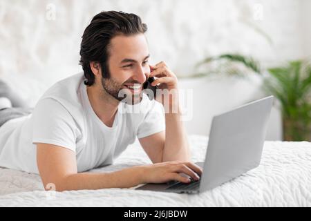 Smiling man making phone call and using laptop in bed Stock Photo
