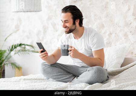 Well-rested handsome guy chilling on bed at home, using smartphone Stock Photo