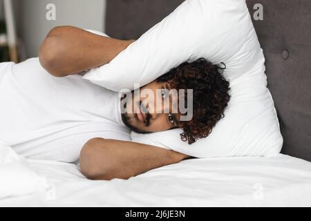 Irritated eastern man laying in bed, covering head with pillow Stock Photo