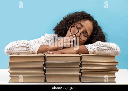 Exhausted young African American woman yawning, lying on big stacks of books, feeling bored while preparing for exam Stock Photo