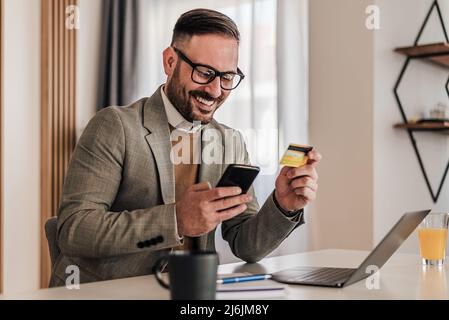 Smiling confident businessman making card payment through smart phone. Happy professional doing online transaction. He is working on laptop at office Stock Photo