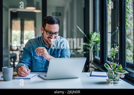 Happy male entrepreneur writing ideas on note pad. Businessman working on laptop at table. He is doing freelance work at home office. Stock Photo