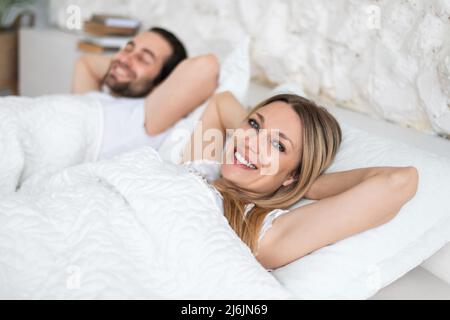 Happy young married couple relaxing on soft pillows in bed, enjoying lazy weekend morning at home, copy space Stock Photo