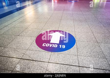 A round signpost with the inscription 'Covid 19' and an arrow is stuck as an indicator on the tiled floor of a public building. Stock Photo