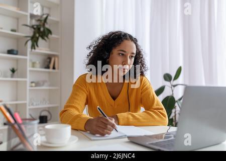 Focused young black woman in headset having online lesson on laptop, writing in notebook at home Stock Photo