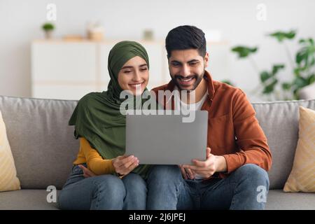 Happy Middle Eastern Spouses Using Laptop Sitting On Couch Indoor Stock Photo