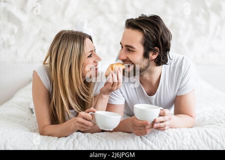 Loving young couple eating breakfast and drinking coffee in bed at home. Lazy romantic morning concept Stock Photo