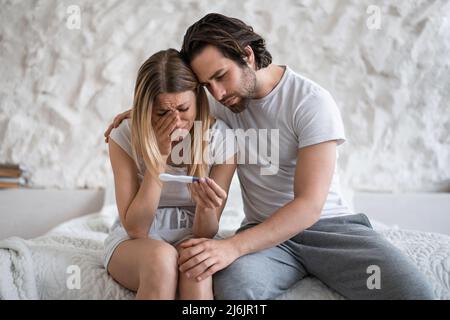 Upset young woman with negative pregnancy test crying on bed, loving husband supporting her at home Stock Photo