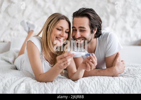 Happy young woman and her husband lying on bed with positive pregnancy test at home Stock Photo