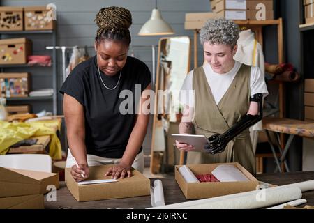 Happy girl with partial arm using tablet while her colleague packing parcel with folded clothes and sticking address of client on its top Stock Photo