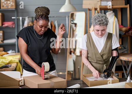 Two young female workers of warehouse packing and sealing with cellotape parcels with packed goods ordered by online clients Stock Photo