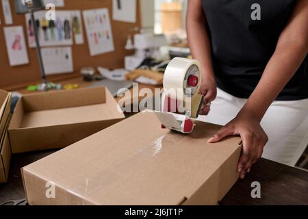 Close-up of African American female worker of warehouse sealing packed cardboard box with client order with cellotape by workplace Stock Photo