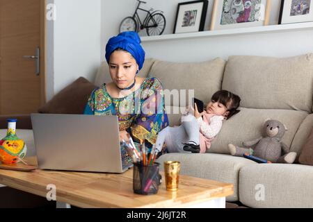 Young Muslim woman working at home with her laptop while her young daughter plays with a mobile phone. Single parent family. Stock Photo